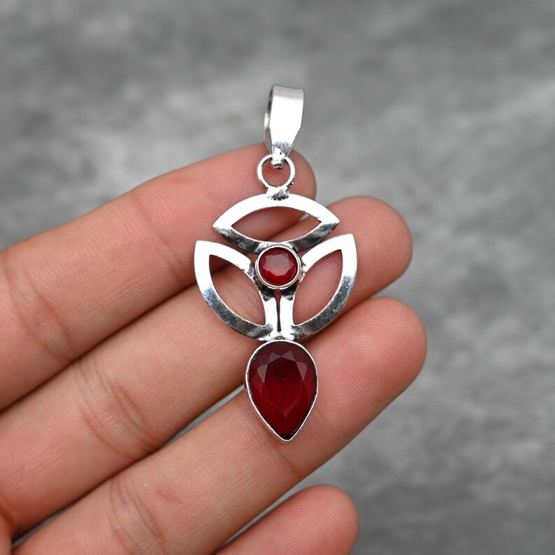Pu&ograve; includere: Un ciondolo d'argento con un design geometrico che presenta due pietre preziose rosse. Il ciondolo ha la forma di un fiore stilizzato con una pietra preziosa a forma di lacrima che pende dal centro.