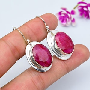 May include: A pair of silver drop earrings featuring large, oval-cut, ruby-red gemstones. The earrings have a simple, elegant design with a polished silver frame around each stone. The gemstones are a vibrant, deep red color.