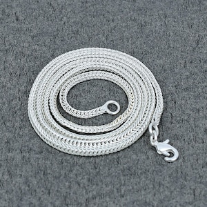 Puede incluir: Un collar de cadena plateada con un diseño tejido, enrollado sobre una superficie gris. El collar tiene un cierre de mosquetón y un pequeño lazo en el otro extremo. La cadena está hecha de pequeños eslabones interconectados.