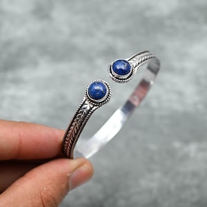 May include: Silver bangle bracelet with two blue lapis lazuli gemstones. The bracelet has a decorative, leaf-like design.