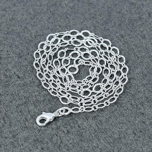 Puede incluir: Un collar plateado con un diseño de cadena de eslabones ovalados. El collar está colocado sobre una superficie gris y presenta un cierre de mosquetón. Los eslabones de la cadena tienen un aspecto texturizado y martillado.