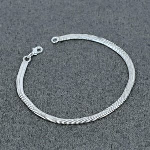 May include: A silver-toned herringbone chain bracelet featuring a flat, flexible link design with a polished metallic finish. The bracelet includes a small lobster claw clasp at one end, allowing for a secure closure when worn around the wrist.