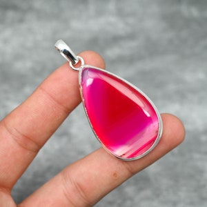 May include: A teardrop-shaped pendant with a vibrant pink and red gemstone set in a silver-colored metal frame. The polished stone displays a layered pattern. The pendant has a loop for a chain.