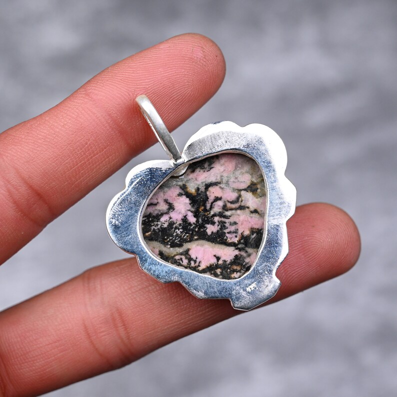 Op de afbeelding: Zilveren hanger met een hartvormige steen met roze en zwarte patronen. De steen is gezet in een decoratief zilveren frame met een lus voor een ketting. De hanger wordt door een vinger vastgehouden.