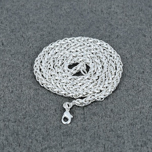 Puede incluir: Un collar de cadena plateada con un diseño de eslabones de trigo, enrollado sobre una superficie gris. El collar tiene un cierre de mosquetón. La cadena es de un material metálico brillante.