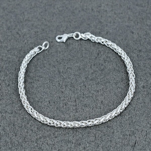 May include: A silver-toned bracelet featuring a continuous woven wheat chain design. The bracelet is constructed with interlocking metal links that create a textured, braided appearance and includes a small lobster claw clasp at one end.