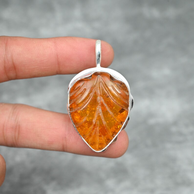 May include: A silver pendant featuring a carved amber leaf. The amber is a warm orange color and has a translucent quality. The pendant is a unique and beautiful piece of jewelry.