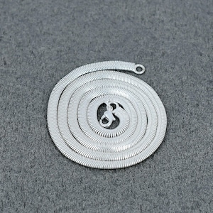 May include: A coiled, silver-coloured necklace with a flat, herringbone chain design. The necklace features a lobster clasp closure and a small loop at the end. The chain is approximately 0.5 cm wide. The necklace is displayed on a grey surface.