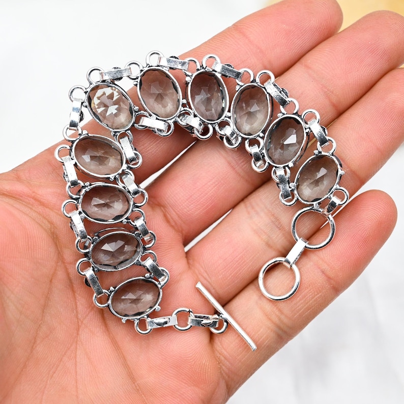 Puede incluir: Un brazalete de plata con piedras preciosas de cuarzo ahumado de forma ovalada. El brazalete tiene un cierre de palanca.