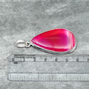 May include: A teardrop-shaped pendant with a vibrant pink and red agate stone set in a silver-colored metal frame. The pendant measures approximately 2.5 inches in length. The stone has a polished, glossy finish.