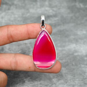 May include: A teardrop-shaped pendant with a vibrant pink and red gemstone set in a silver-colored metal frame. The gemstone has a layered appearance, with bands of color. The pendant is suspended from a silver bail.