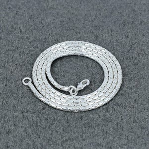 May include: A silver necklace with a snake chain pattern. The chain is coiled on a grey surface, with a clasp and a loop at the ends. The chain is made of small, interlocking metal pieces.