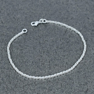 Puede incluir: Una delicada pulsera de cadena de plata que presenta una serie de eslabones planos entrelazados en forma de chevrón. La pulsera termina con una pequeña anilla circular en un extremo y un cierre de mosquetón en el otro para un cierre seguro.