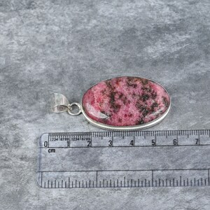 May include: A silver pendant with a pink and black rhodochrosite stone. The stone is oval shaped and measures approximately 2.5 cm in length. The pendant is on a white surface with a ruler for scale.