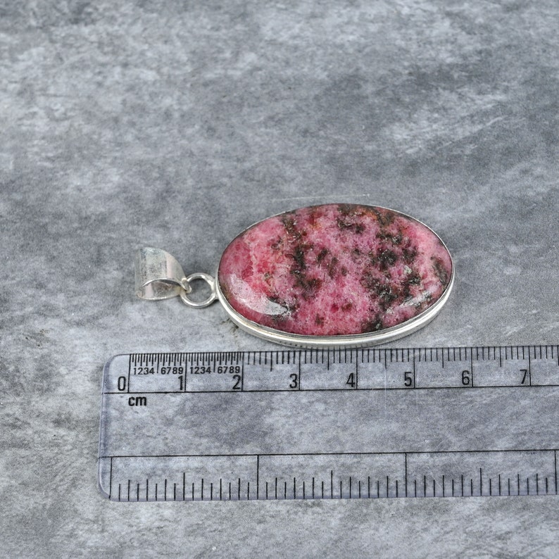 May include: A silver pendant with a pink and black rhodochrosite stone. The stone is oval shaped and measures approximately 2.5 cm in length. The pendant is on a white surface with a ruler for scale.