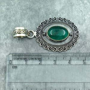 May include: Oval-shaped pendant with a vibrant green gemstone set in a silver-toned frame. The frame features an ornate, swirling design. The pendant includes a decorative bail for attaching to a chain. The ruler shows the pendant is approximately 1 inch in length.
