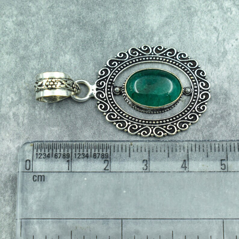 May include: Oval-shaped pendant with a vibrant green gemstone set in a silver-toned frame. The frame features an ornate, swirling design. The pendant includes a decorative bail for attaching to a chain. The ruler shows the pendant is approximately 1 inch in length.