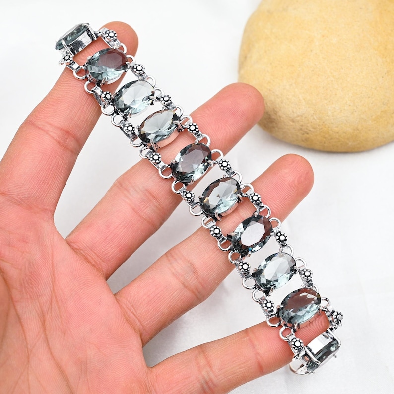 Puede incluir: Un brazalete de plata con un dise&ntilde;o de cadena y piedras preciosas grises de forma ovalada.