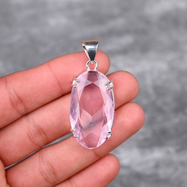 May include: A pale pink oval-shaped gemstone pendant with a silver setting. The pendant is held in a hand.