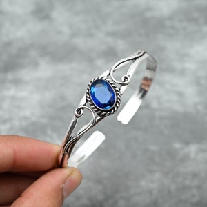 Pode incluir: Uma pulseira de punho prateada com uma grande pedra preciosa oval azul escuro. A pedra preciosa está engastada numa moldura decorativa em forma de corda. A pulseira tem um design aberto com detalhes elegantes e giratórios.