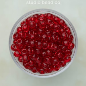 May include: A circular container filled with small, translucent, deep red glass seed beads. The text 'studio bead co' is printed in white at the top center of the image. The beads are uniform in shape and size, creating a vibrant, monochromatic display of red.