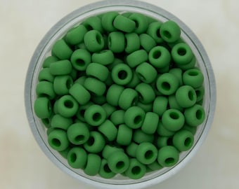 May include: A clear circular container filled with numerous small, matte, forest green pony beads. The text "studio bead co" is printed in white at the top center of the image. The beads are uniform in shape, featuring a central hole for threading.