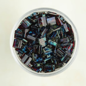 May include: A close-up view of a clear glass jar filled with dark blue and red iridescent glass beads. The beads are rectangular in shape and have a mottled, textured surface.