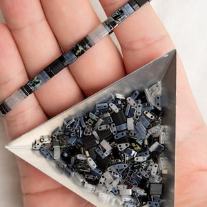 Op de afbeelding: Een driehoekige schaal gevuld met zwarte, grijze en witte glazen kralen. De kralen zijn in verschillende vormen en maten. Een hand houdt de schaal vast en een streng van vergelijkbare kralen.