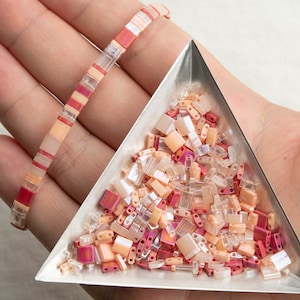May include: A close-up of a triangular tray filled with a variety of small, colorful glass beads. The beads are in shades of pink, orange, and clear. A hand is holding a bracelet made of similar beads in the background.