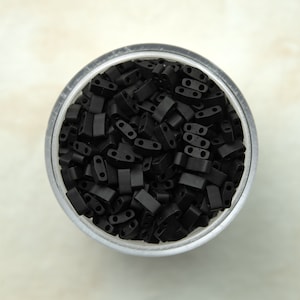 Puede incluir: Un recipiente de plástico transparente lleno de cuentas de plástico negras. Las cuentas son pequeñas y rectangulares con dos agujeros en cada una.