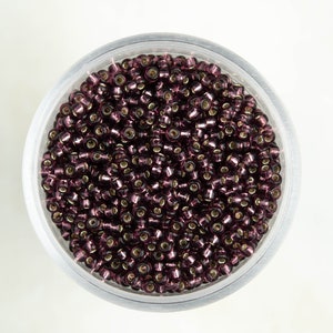 May include: A clear plastic container filled with small, round, iridescent purple glass beads.