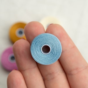 Peut inclure: Une main tenant une bobine de fil bleu clair. La bobine est enroul&eacute;e serr&eacute;e et a un petit trou au centre. Il y a d'autres bobines de fil en arri&egrave;re-plan, y compris du jaune, du rose et du blanc.
