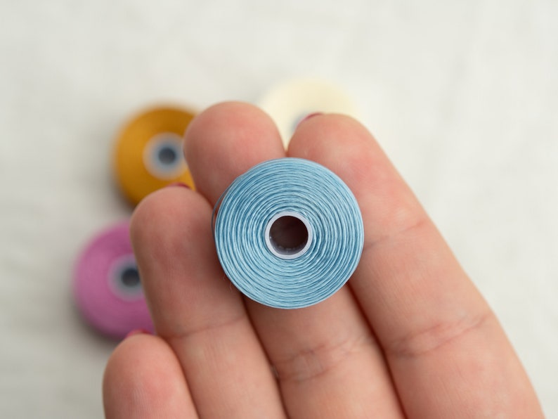 Peut inclure: Une main tenant une bobine de fil bleu clair. La bobine est enroul&eacute;e serr&eacute;e et a un petit trou au centre. Il y a d'autres bobines de fil en arri&egrave;re-plan, y compris du jaune, du rose et du blanc.