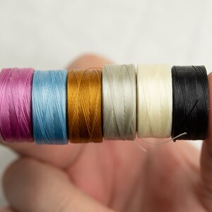 Peut inclure: Six bobines de fil de diff&eacute;rentes couleurs, dont rose, bleu, or, gris, blanc et noir. Les bobines sont tenues dans une main.