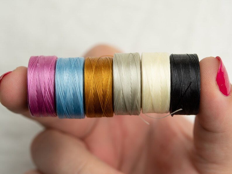 Peut inclure: Six bobines de fil de diff&eacute;rentes couleurs, dont rose, bleu, or, gris, blanc et noir. Les bobines sont tenues dans une main.