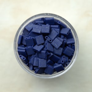 May include: A clear plastic container filled with dark blue square-shaped beads.
