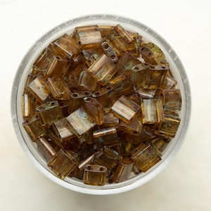 Peut inclure: Un bol rempli de petites perles de verre brunes et irisées. Les perles sont de forme rectangulaire et ont une surface légèrement texturée.