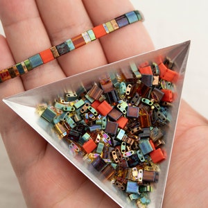 Può includere: Primo piano di un vassoio triangolare pieno di perline di vetro colorate. Le perline sono un mix di arancione, marrone, verde e oro. Il vassoio è appoggiato su una mano, e un braccialetto fatto di perline simili è visibile sullo sfondo.