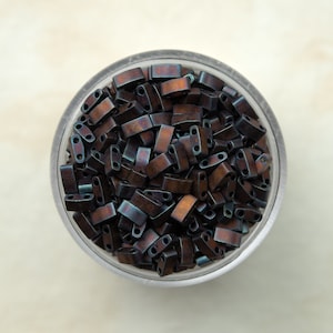 Puede incluir: Un tarro de vidrio transparente lleno de cuentas rectangulares de color marrón oscuro e iridiscente azul.