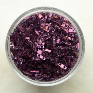 May include: A bowl filled with small, iridescent purple glass beads. The beads are shaped like elongated rectangles with two holes in each.