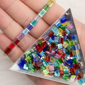 May include: A close-up of a triangular tray filled with colorful iridescent glass beads. The beads are in various shapes and sizes, with a rainbow of colors including red, blue, green, yellow, and purple. The beads are arranged in a random pattern, creating a visually appealing display.