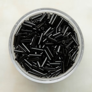 Op de afbeelding: Een glazen pot gevuld met zwarte glazen kralen. De kralen zijn lang en cilindrisch, en lijken op kleine buisjes.