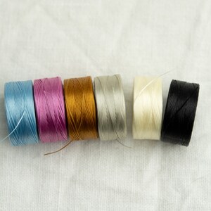 Peut inclure: Six bobines de fil de diff&eacute;rentes couleurs, dont bleu clair, rose, or, gris, blanc et noir. Les bobines sont dispos&eacute;es en rang&eacute;e sur une surface blanche.