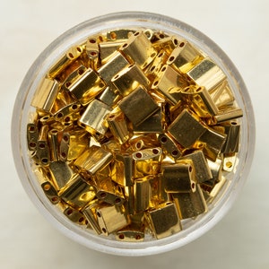 May include: A close-up of a glass jar filled with gold-coloured metal beads. The beads are small and rectangular with three holes in each.