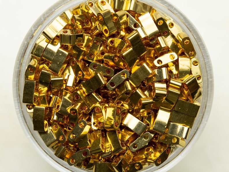 Może przedstawiać: Widok z bliska pojemnika wypełnionego złotymi metalowymi k&oacute;łkami do łączenia. K&oacute;łka są małe i mają prostokątny kształt.
