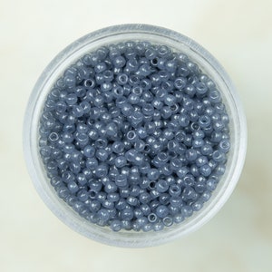 May include: A transparent container filled with many small, round, blue-grey beads. The beads are consistent in size and shape, and appear to be made of glass or a similar material. The container is set against a light-coloured background.