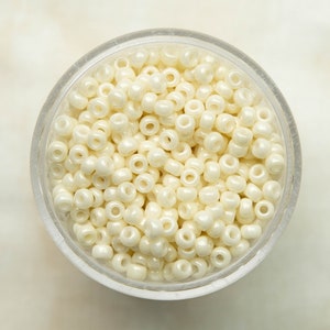 May include: A close-up view of a small clear container filled with white seed beads. The beads are round and have a slight sheen.