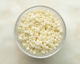 May include: A close-up view of a small clear container filled with white seed beads. The beads are round and have a slight sheen.
