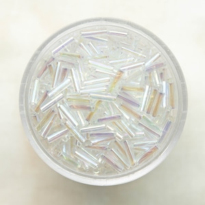May include: A close-up view of a bowl filled with iridescent glass beads. The beads are small and cylindrical, with a clear, translucent appearance. The beads have a subtle rainbow sheen.