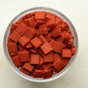Può includere: Una ciotola piena di piccole perline quadrate di colore rosso-arancio. Le perline hanno tre fori in ogni angolo.